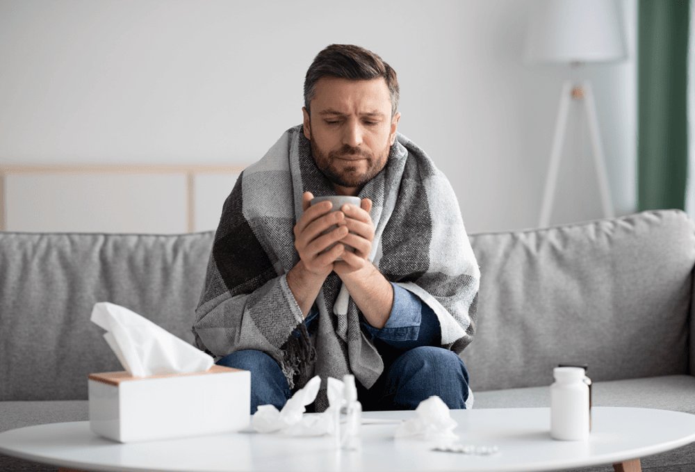 Man with the flu holding a warm drink with a blanket round him